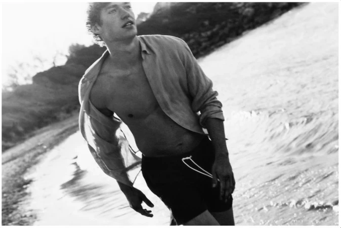 A man with wavy hair wears an unbuttoned shirt and dark swim trunks while walking along a beach shoreline in a black and white photo.