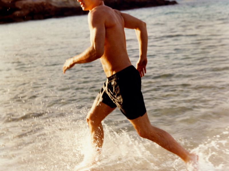 An adult male runs through shallow ocean water at the beach on a sunny day, captured at eye-level with a focus on motion and the surrounding environment.