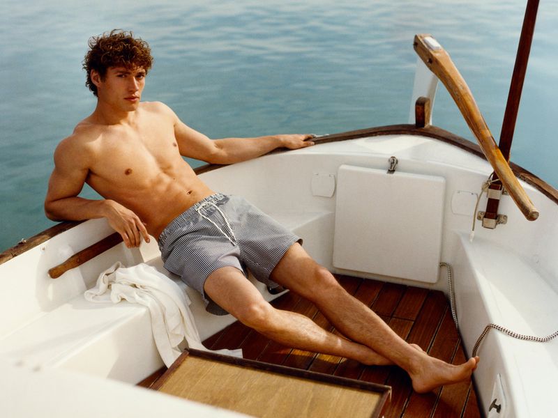 Shirtless man in striped swim trunks reclines on a boat deck against blue water on a sunny day; relaxation and summer vibes.