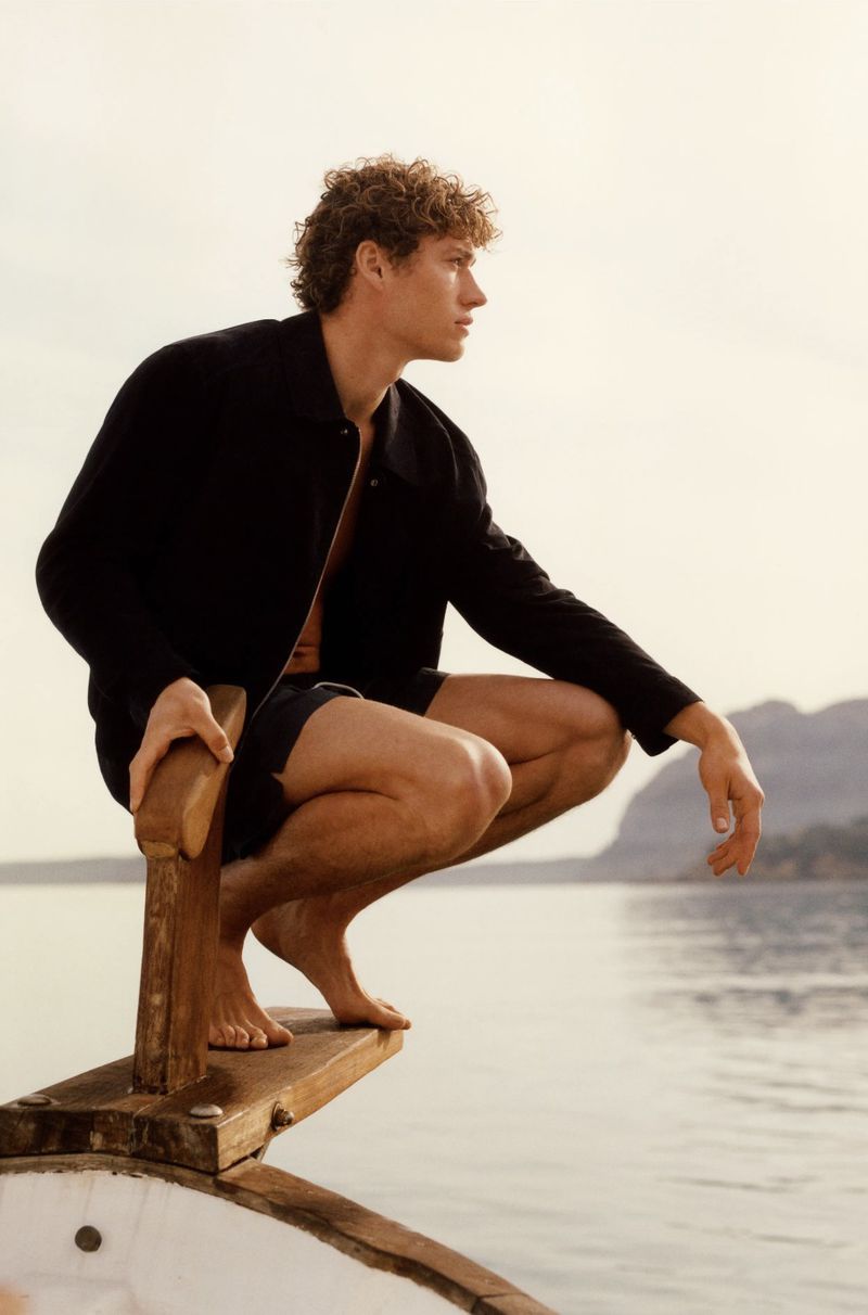 Man in dark jacket crouches on a boat overlooking ocean and distant land, captured in a medium shot under bright daylight.