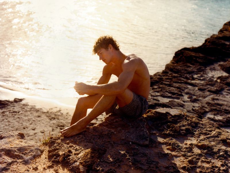 Young adult male sits shirtless on rocks at beach; sun highlights tousled hair and ocean backdrop, creating a pensive, summery portrait.