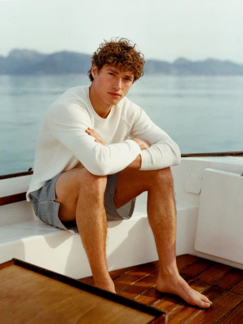 A curly-haired man in a white sweatshirt and striped shorts sits barefoot on a boat, looking at the camera, with water and islands behind.