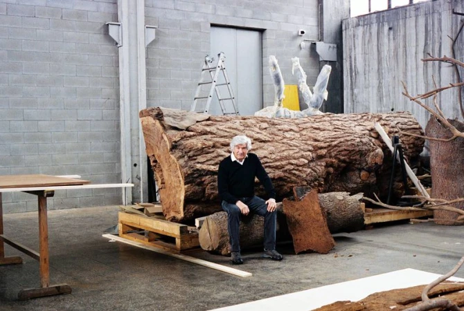 Mature man sits in front of a huge tree trunk in an industrial setting, next to a ladder, with cement walls and an elevator in background.