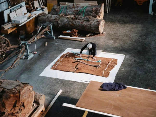An adult uses measuring tape to measure a piece of bark on a white surface in a workshop; trees and tools are visible in background.
