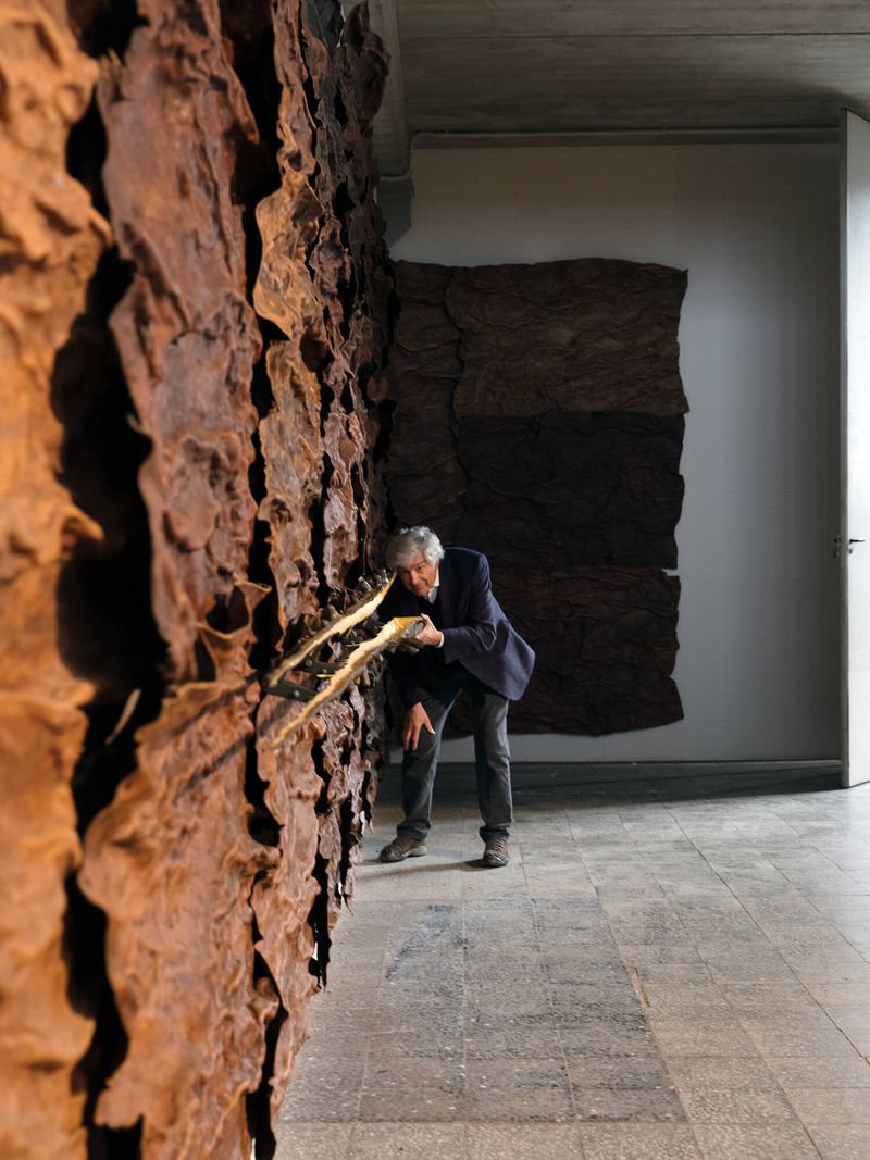 Adult male in a suit crouches to examine a textured brown wall in an art gallery under bright light, with a minimalist aesthetic.