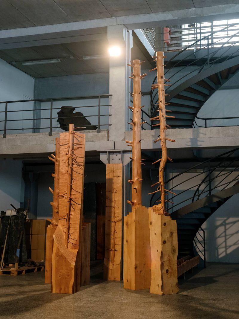 Wooden tree sculptures stand in an industrial space near a spiral staircase, artfully crafted for an indoor display.