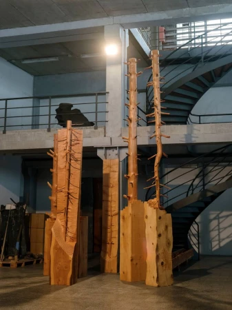 Wooden tree sculptures stand in an industrial space near a spiral staircase, artfully crafted for an indoor display.