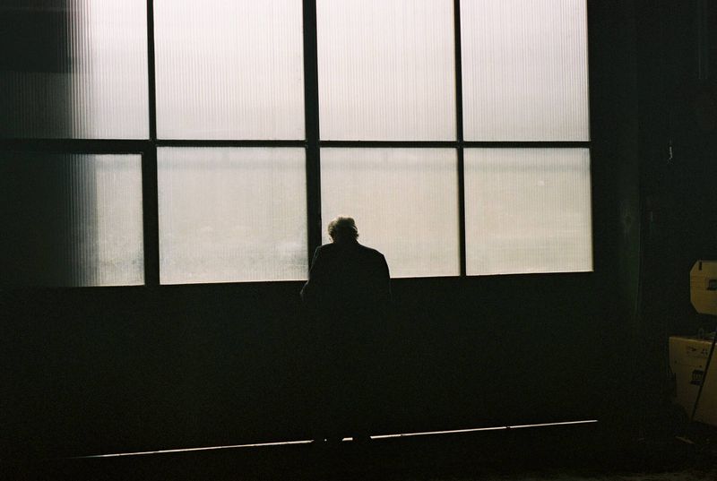 A silhouetted person stands before a large, divided window in a dark industrial space, creating a contrast between light and shadow.