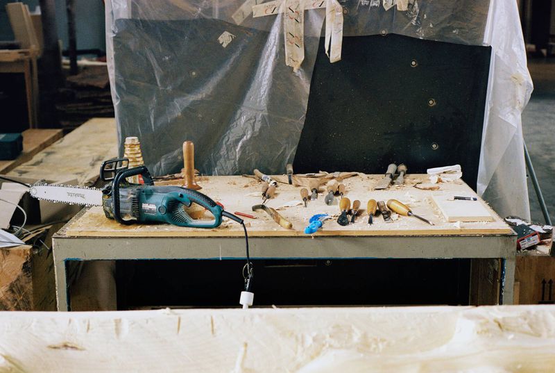Workshop table with a chainsaw and carving tools under a plastic tarp, creating an atmosphere of craftsmanship in an indoor setting.