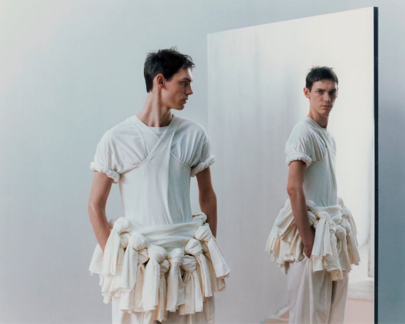 Man posing in white outfit next to a mirror reflecting his image. Studio shot with bright lighting and neutral background.