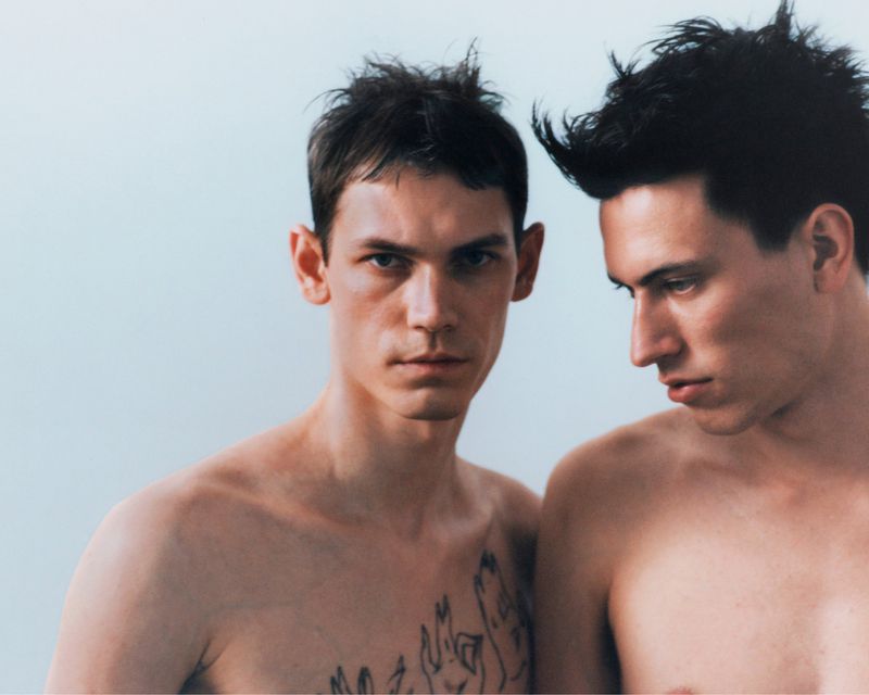 Two shirtless men posing against a pale studio background with dark hair and chest tattoos