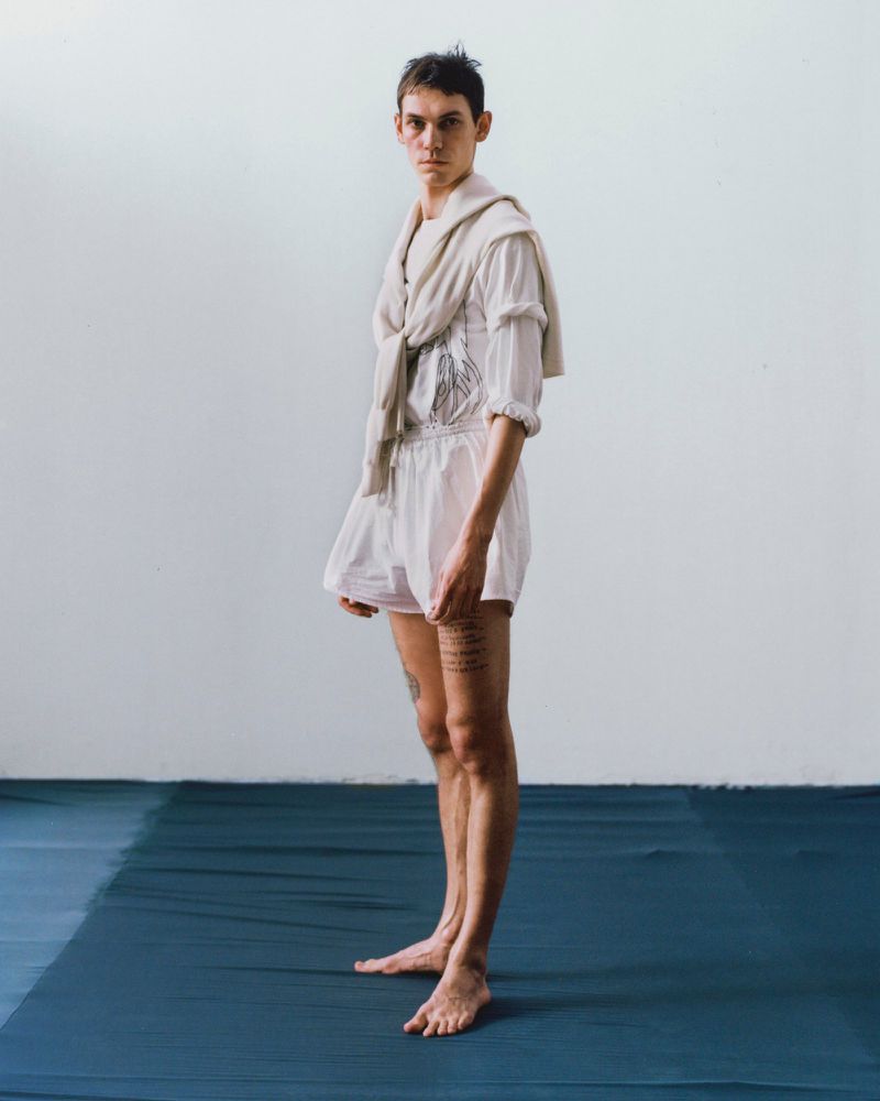 Full-length portrait of a slender young adult, fair-skinned, in a light-colored outfit, posed against a neutral background in a photo studio.