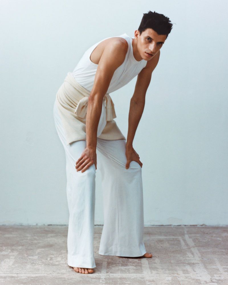 A man in a white tank top and wide-legged pants leans forward with hands on his knees in a studio setting against a white wall.