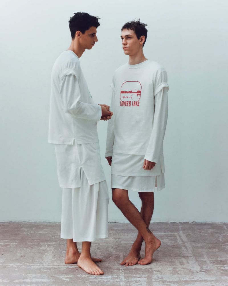 Two young adult male fashion models pose indoors wearing oversized, layered white shirts and shorts against a neutral background.