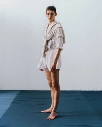 A barefoot male model posing in white shorts and a draped off-white sweater against a plain white wall and blue floor.