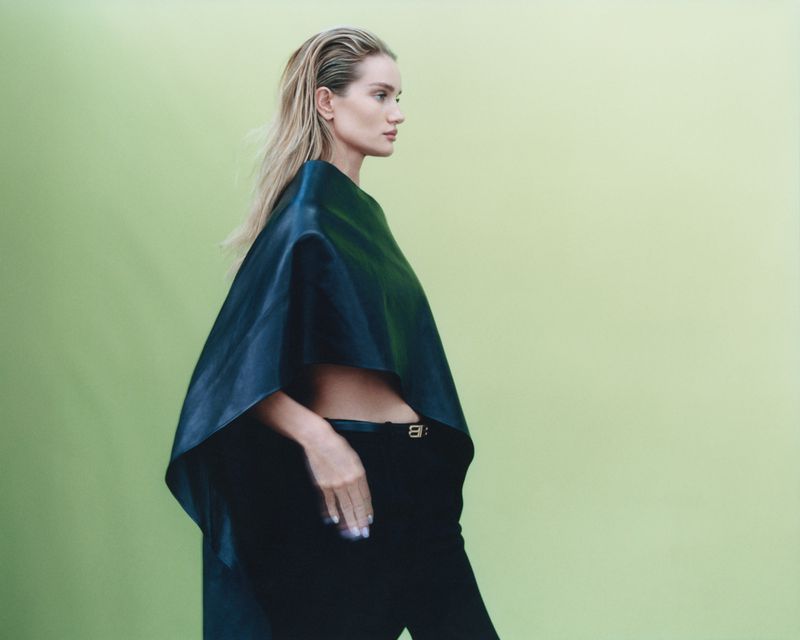 Blonde woman in black pants and top poses against a green background in a fashion style photograph with studio lighting.