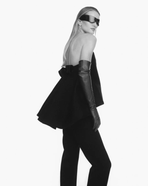 Monochrome fashion shot of a woman in a black, backless top, trousers, opera gloves, and visor sunglasses against a white background.