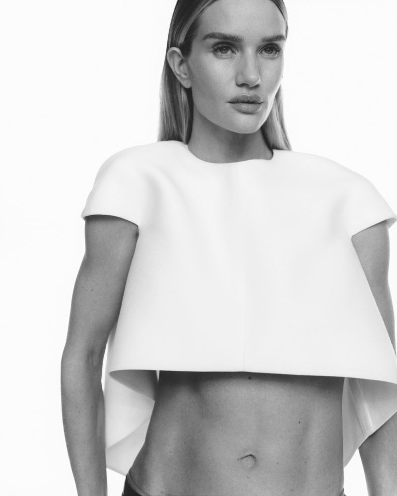 Black-and-white portrait of a woman in a cropped white top against a stark white backdrop. The woman is posing with a neutral expression.