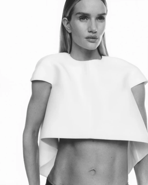 Black-and-white portrait of a woman in a cropped white top against a stark white backdrop. The woman is posing with a neutral expression.