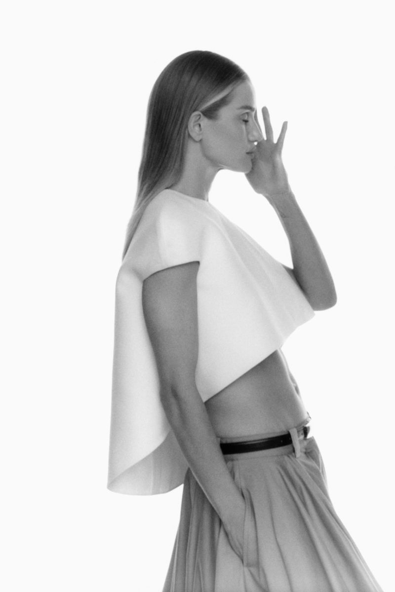 Black and white portrait of a slender woman in cropped top and skirt. She is holding her hand to her face with a neutral expression.