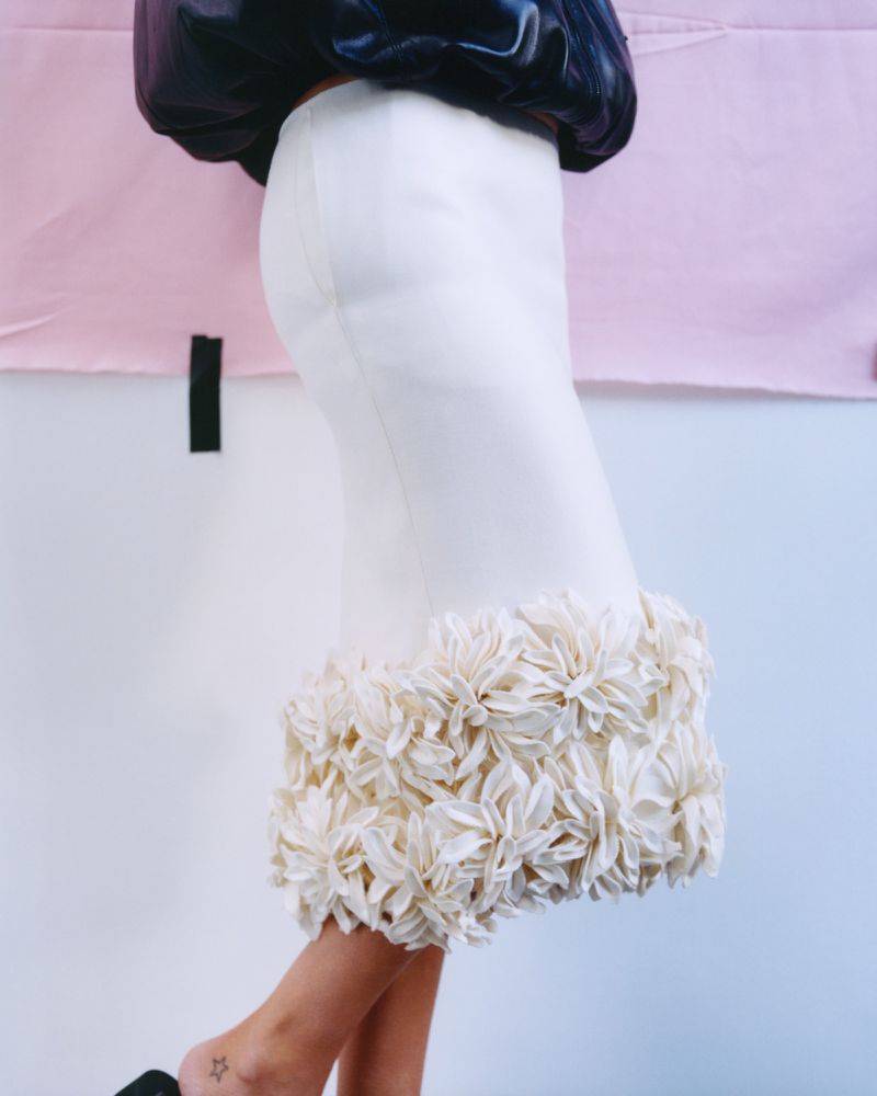 Close-up of a woman's lower torso wearing a white pencil skirt with a floral ruffle hemline, paired with black shoes and a black top.