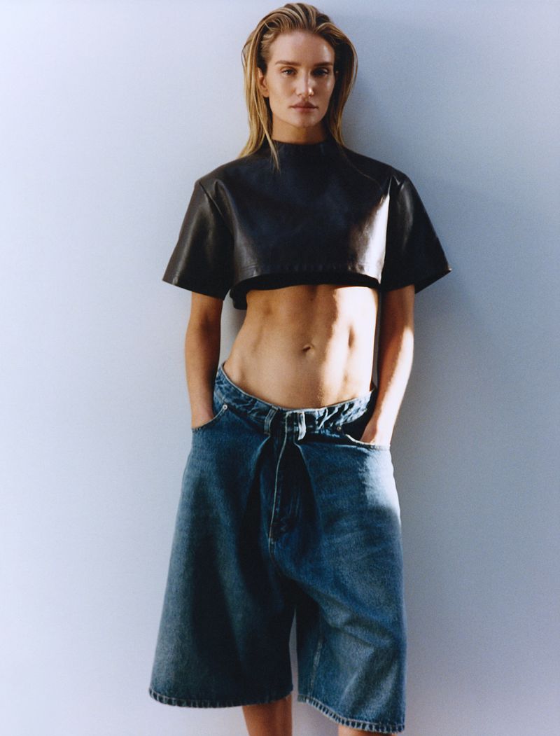 A stylish woman in a cropped black top and baggy denim shorts poses against a grey backdrop in a fashion editorial setting.