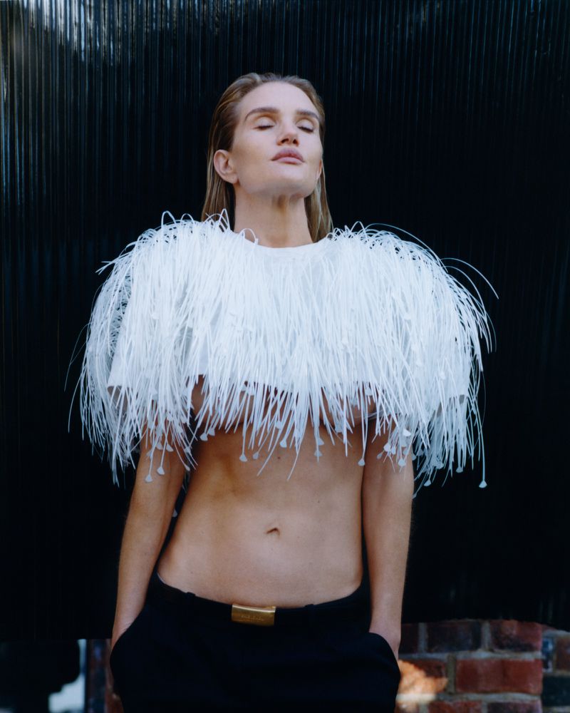 Fashionable adult female model poses with fringed couture top and black pants against a black wall with partial brick detail.