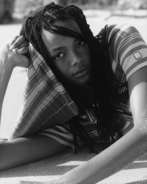 Black and white portrait of a woman with dreadlocks laying on a beach with a striped shirt, eyes looking directly at the camera.