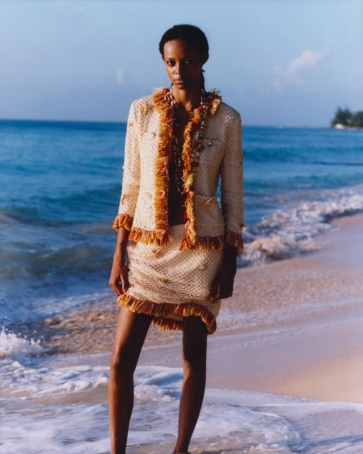 A woman in a tweed jacket and skirt stands on a sandy beach, with the ocean in the background, in this outdoor fashion shot.