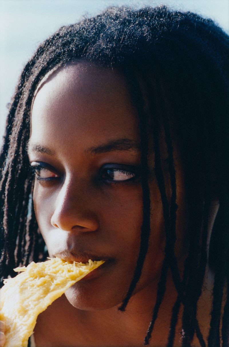 Close-up of a Black woman with dreadlocks eating a snack outside, with natural lighting and a shallow depth of field.