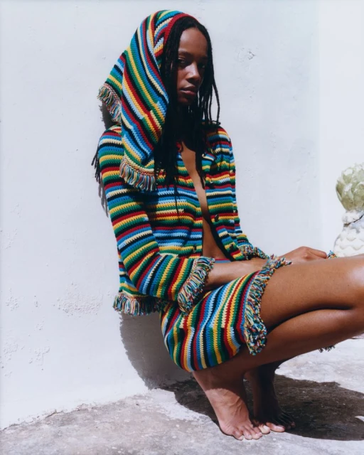 Adult female crouches in a colorful, fringed, striped hooded jacket and skirt, gazing to the right against a minimalist backdrop. Fashion shot.