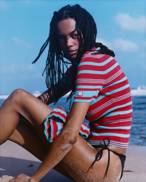 Young woman with dark dreadlocks wearing a striped top and bikini bottoms sits on a sandy beach near the ocean on a sunny day.