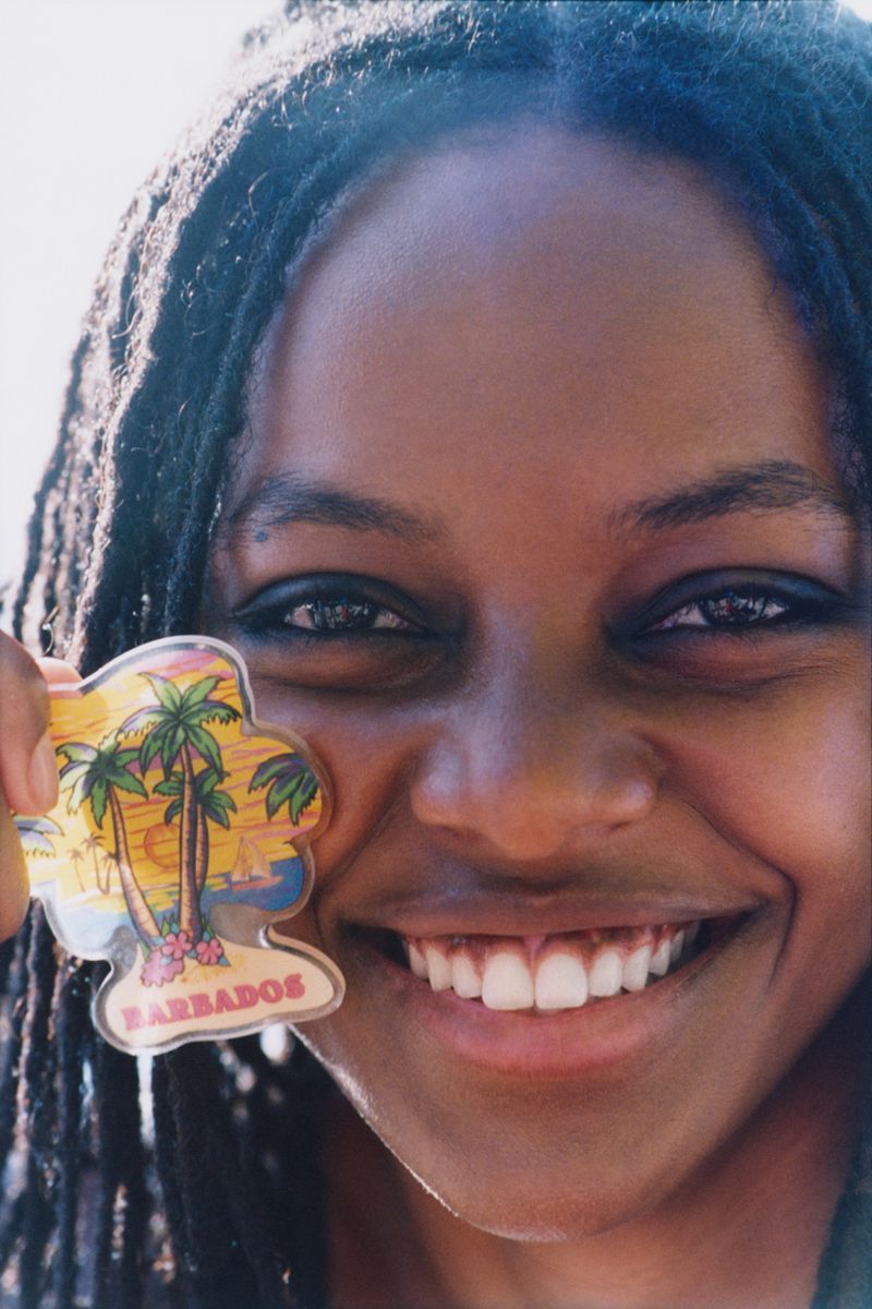 Close-up portrait of smiling woman with dreadlocks, holding a Barbados souvenir magnet with palm trees and a beach scene.