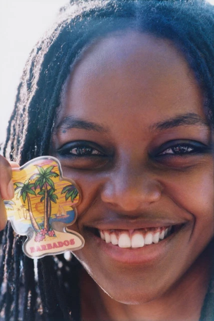 Close-up portrait of smiling woman with dreadlocks, holding a Barbados souvenir magnet with palm trees and a beach scene.