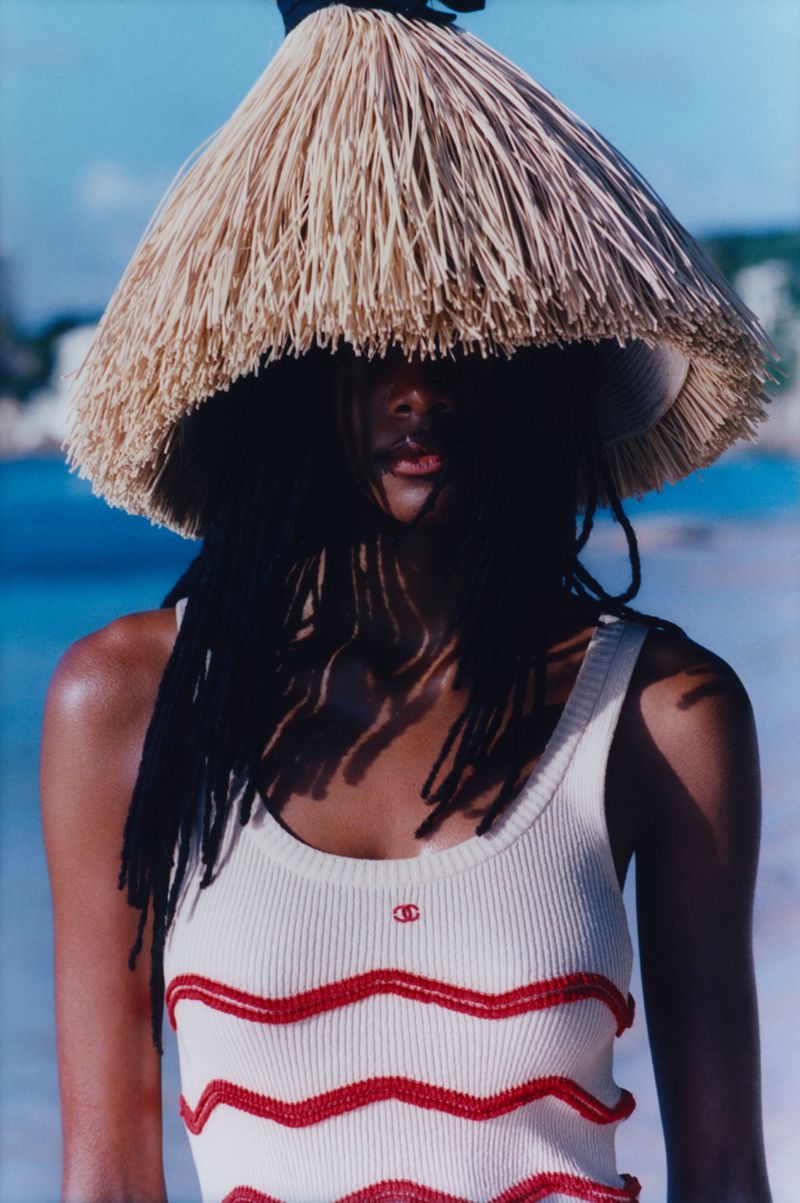 Portrait of a Black woman in a white tank top with a straw hat, standing outside with ocean and vegetation in the background on a sunny day.