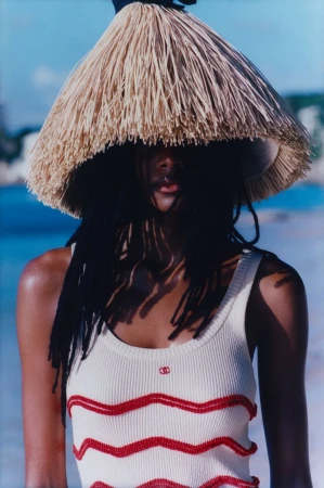 Portrait of a Black woman in a white tank top with a straw hat, standing outside with ocean and vegetation in the background on a sunny day.
