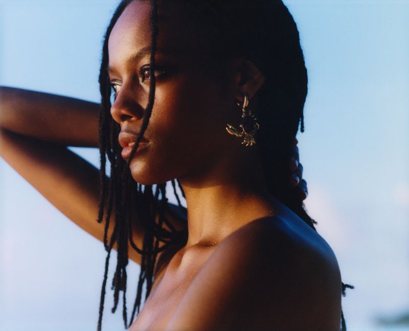 Close-up portrait of a young adult woman with dark hair and dark skin wearing a gold earring against a blurred blue sky background.