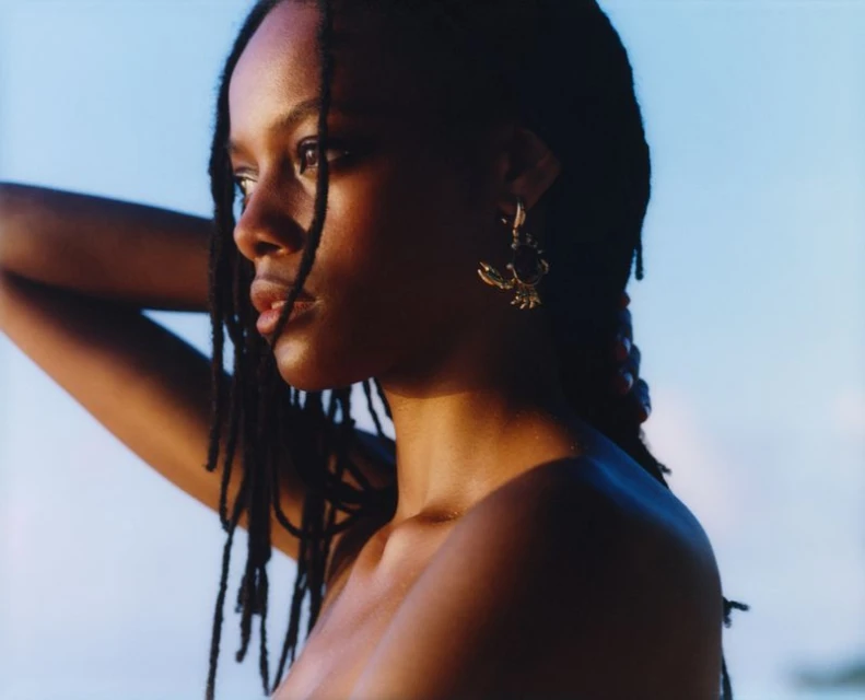 Close-up portrait of a young adult woman with dark hair and dark skin wearing a gold earring against a blurred blue sky background.