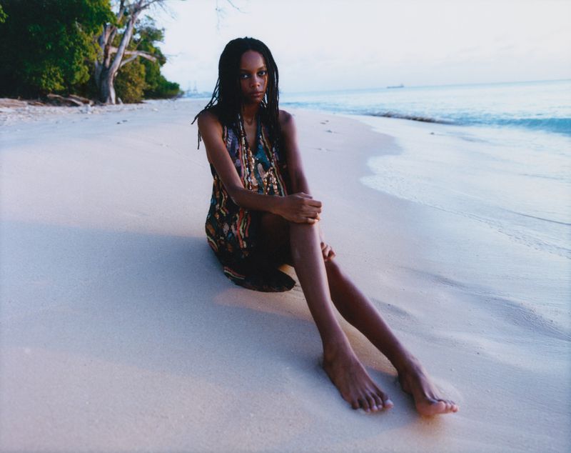 A woman with long dreadlocks sitting on a sandy beach near the ocean wearing a patterned dress, posing for a fashion-style photo.