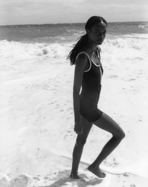 A young woman walks along a black and white beach; she wears a retro one-piece swimsuit as she walks through the surf on a sunny day.