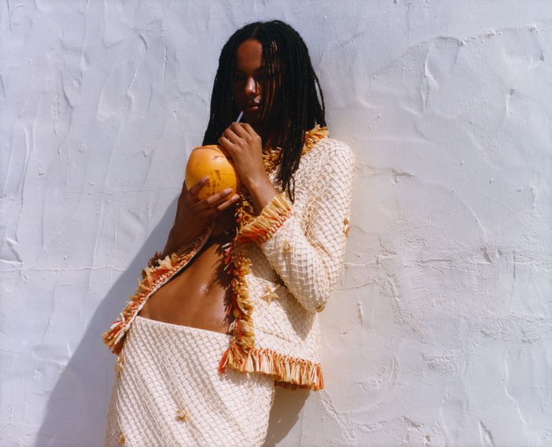A woman in a knit suit drinks from a coconut. She poses in front of a white textured wall in sunlight, creating a relaxed fashion shot.