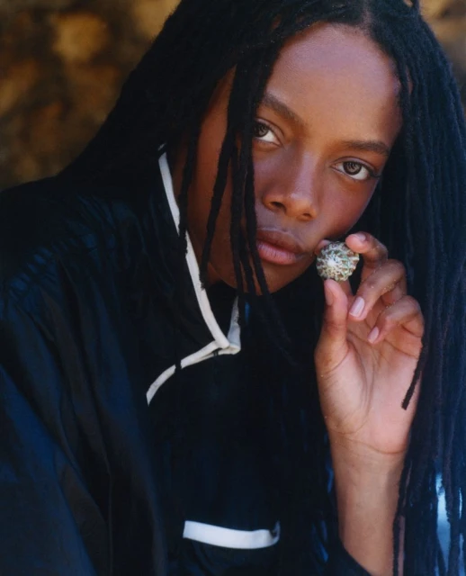 Portrait of an adult female with dark skin and long dreadlocks, holding a spiky round object, wearing a black jacket.