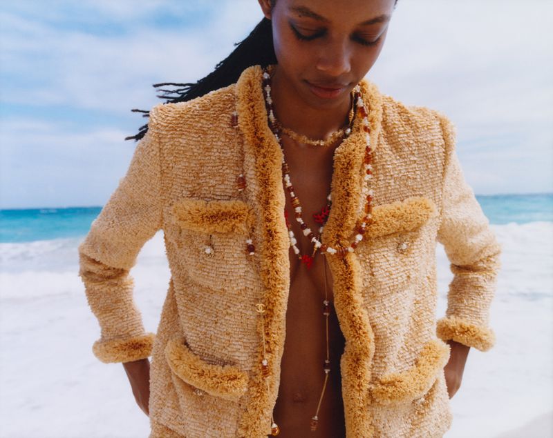 A woman stands on a sandy beach with a textured jacket and beaded necklaces in front of a bright turquoise ocean with small waves.