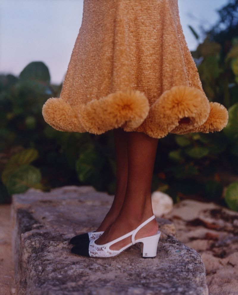 Fashionable woman showcases Chanel slingback heels with a textured beige dress while standing on a stone surface outdoors.