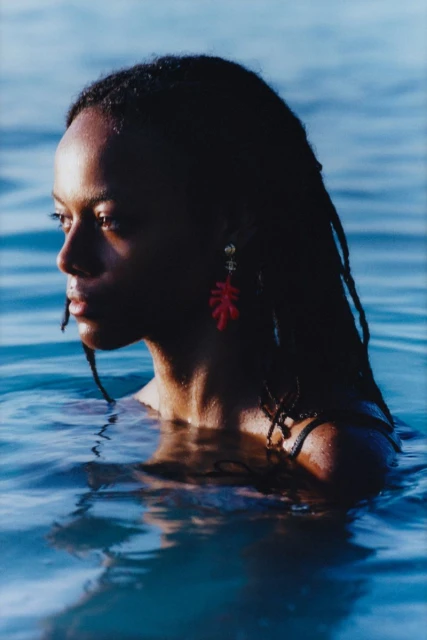 Portrait of a Black woman in ocean water, captured in a medium shot with natural lighting, featuring red coral earrings and a serene expression.
