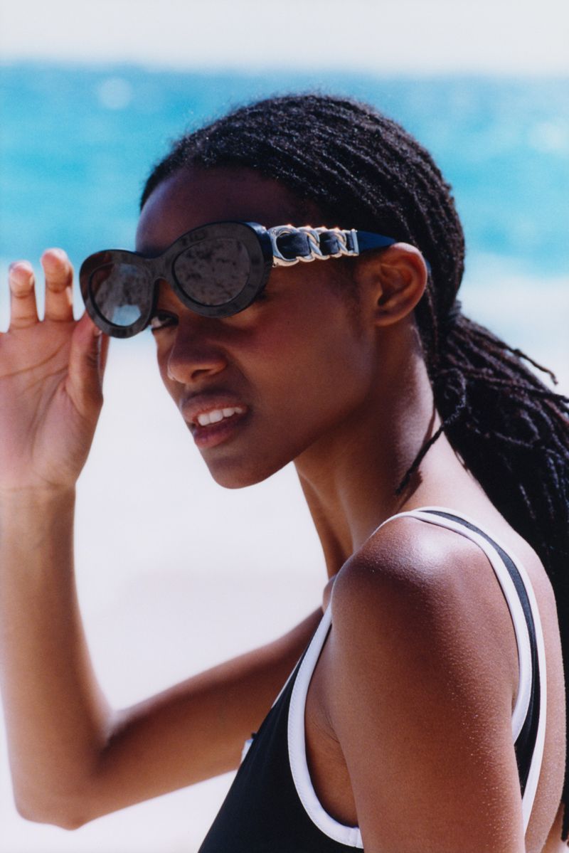 Headshot of a smiling Black woman with sunglasses posing on a beach with white sand and blue ocean water in the background.