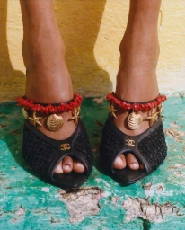Close-up of model Nyela Hopster wearing black Chanel open-toe mesh mules detailed with red beads and dangling gold shell charms.