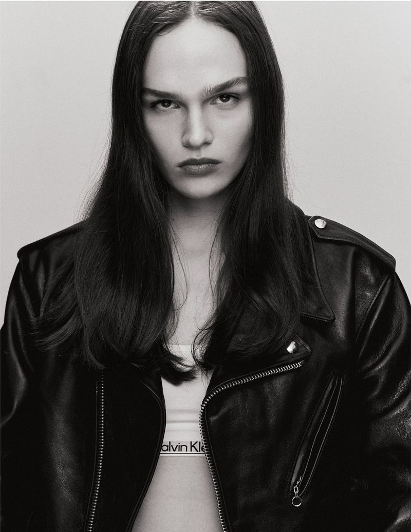 Close-up studio shot of a young adult woman with long dark hair wearing a black leather jacket and white Calvin Klein top, looking at the camera.