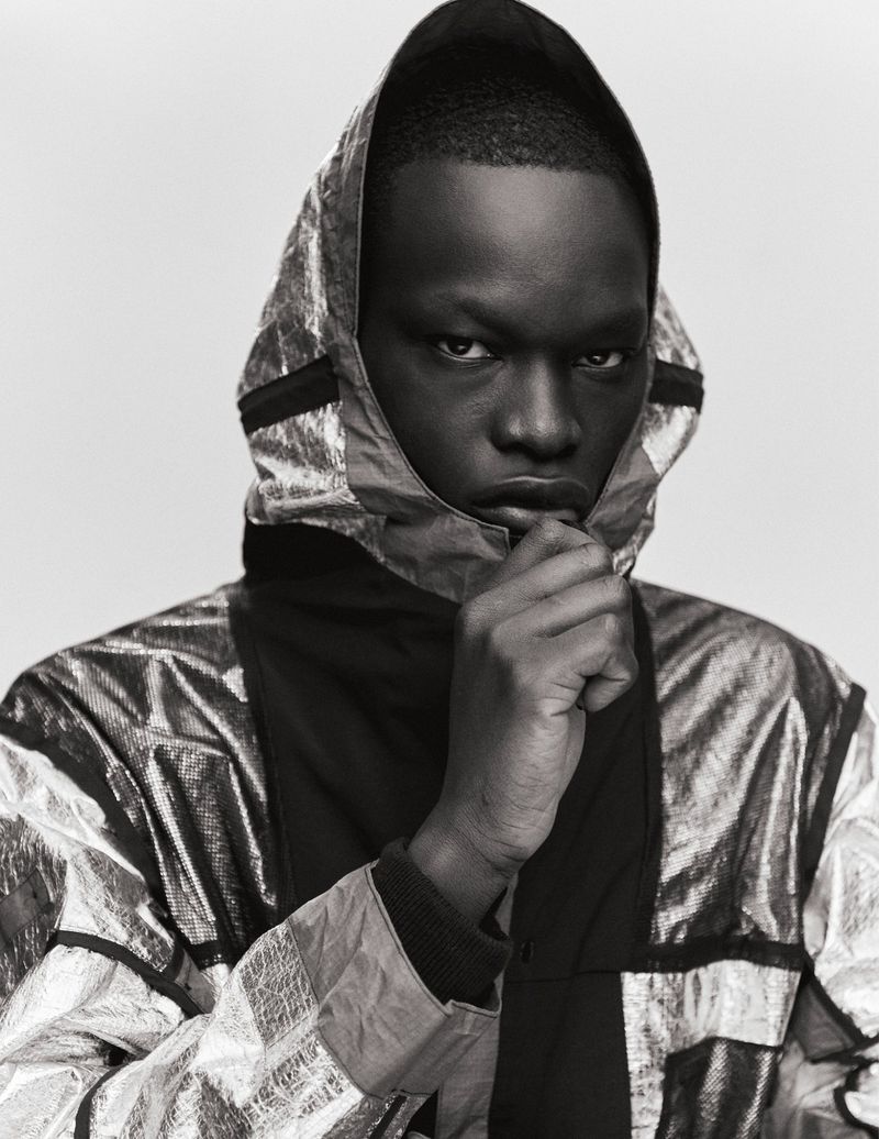 Close-up studio portrait of a Black man with dark skin wearing a metallic silver hooded jacket and looking at the camera.