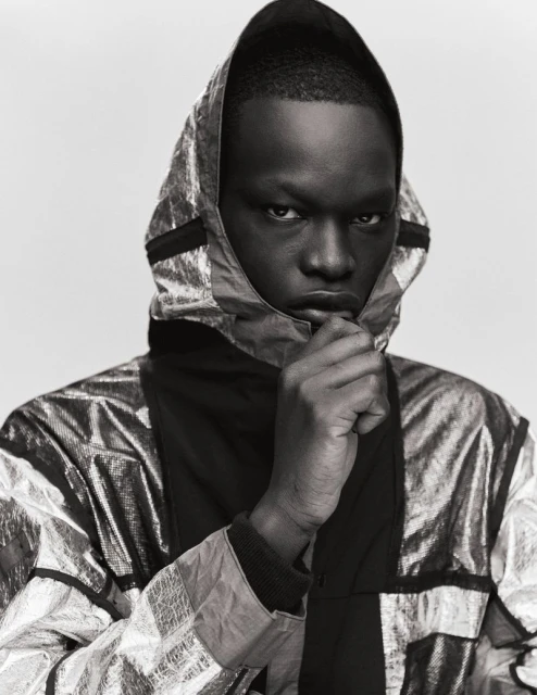 Close-up studio portrait of a Black man with dark skin wearing a metallic silver hooded jacket and looking at the camera.